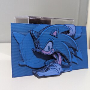 May include: A blue and purple papercraft of Sonic the Hedgehog, a popular video game character, running with his signature blue quills and red shoes. The papercraft is layered and detailed, creating a 3D effect.