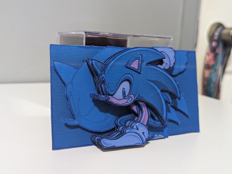 May include: A blue and purple papercraft of Sonic the Hedgehog, a popular video game character, running with his signature blue quills and red shoes. The papercraft is layered and detailed, creating a 3D effect.