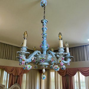 May include: A vintage chandelier with a light blue finish and ornate floral detailing. The chandelier features multiple arms with candle-style light fixtures and is suspended from a chain. The floral accents include pink, blue, and purple flowers.