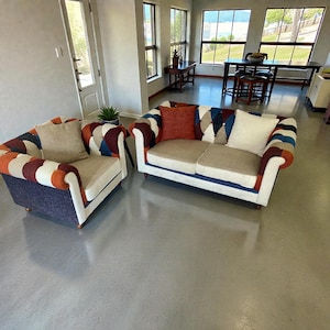 Könnte beinhalten: Ein Wohnzimmerset mit einem gemusterten Sessel und einem Zweisitzer-Sofa. Die Möbel haben ein Patchwork-Design mit verschiedenen Farben, darunter Orange, Blau und Weiß. Beige Kissen und eine blaue Basis vervollständigen den Look. Der Raum hat große Fenster.