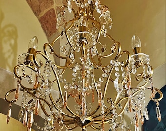 May include: A gold-toned chandelier with multiple arms and crystal accents. The ornate fixture features several light bulbs and cascading crystal droplets, creating a luxurious and elegant aesthetic. The chandelier is designed for indoor use.