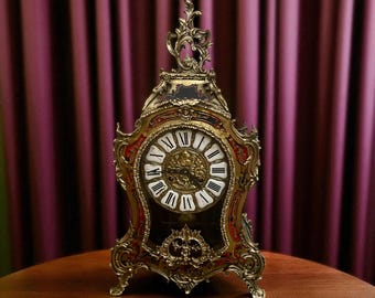 May include: An ornate antique clock with a gold-colored frame and red accents. The clock face features Roman numerals and decorative detailing. The clock is displayed on a wooden surface against a purple curtain backdrop.