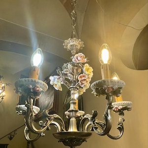 May include: An ornate chandelier with a brass finish, featuring delicate porcelain flowers in shades of pink, yellow, and white. The chandelier has three arms with candle-shaped lights and decorative accents, suspended from a chain.