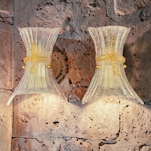 May include: Two clear glass wall sconces with a textured, ribbed design. Each sconce has a gold-colored band and accents. The sconces are mounted on a textured, reddish-brown brick wall, creating a warm, inviting ambiance.