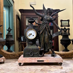 Peut inclure: Horloge ancienne en bronze avec une statue d'une figure ailée, flanquée de deux urnes décoratives sur des socles en marbre. L'horloge a un cadran blanc avec des chiffres romains noirs. Les objets sont exposés sur une table en marbre.