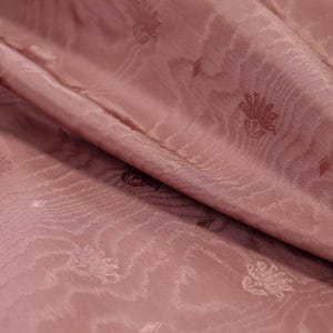 May include: A dusty rose fabric featuring a subtle, repeating floral motif and a wavy, water-like texture woven into the material. The textile is draped in soft folds, highlighting the sheen and the intricate, small-scale pattern across the surface.
