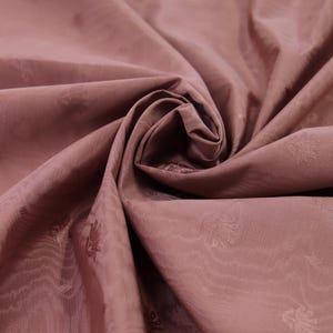 May include: A dusty rose fabric featuring a subtle, repeating tonal floral pattern. The material is gathered in the center, creating soft folds and shadows that highlight the smooth texture and the delicate, woven botanical motifs throughout.