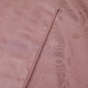 May include: A dusty rose fabric featuring a subtle, tonal jacquard pattern of small, repeating floral motifs. The material has a soft, semi-sheer quality with a visible weave and a slightly textured surface, showing a folded edge.