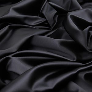 Può includere: Un pezzo di tessuto nero a tinta unita con una finitura liscia e lucente. Il materiale è drappeggiato in pieghe profonde e fluide che creano una serie di luci e ombre sulla superficie, enfatizzando la qualità riflettente del tessuto scuro.