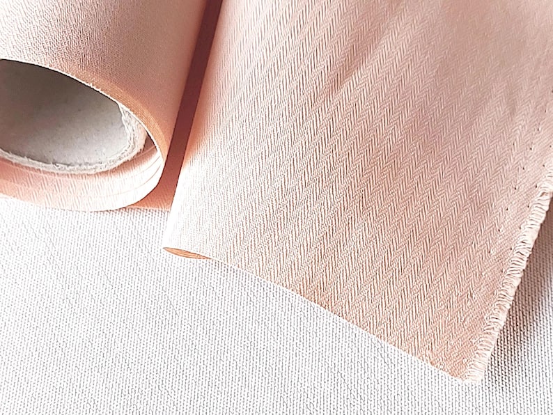 May include: A roll of pale pink fabric with a subtle herringbone pattern. The fabric is soft and has a slightly textured surface.