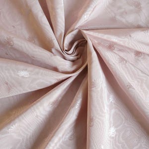 May include: A light pink fabric featuring a subtle, repeating floral pattern and a wavy, moir&eacute;-like texture. The material is gathered in the center, creating deep folds and shadows that highlight the sheen and delicate design of the textile.