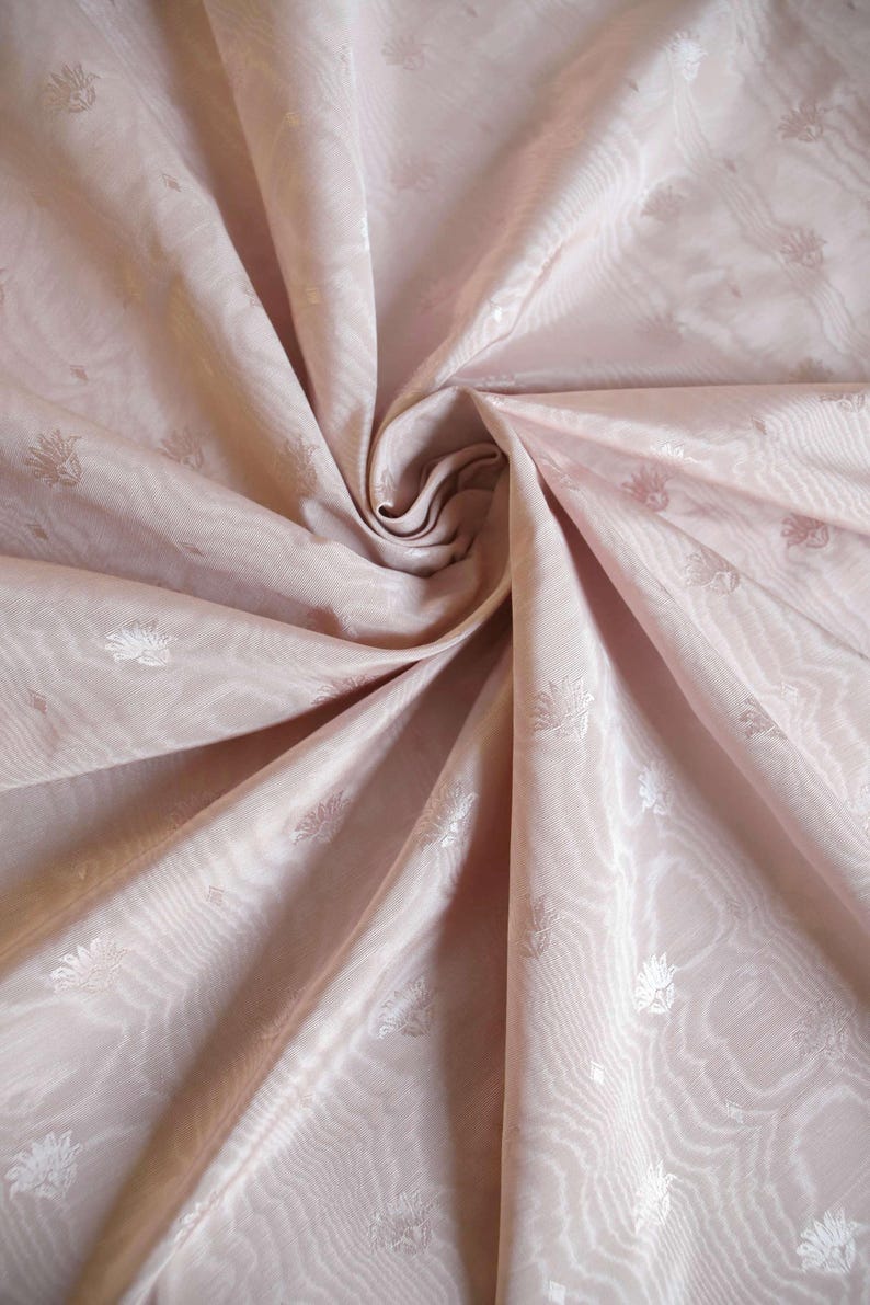 May include: A light pink fabric featuring a subtle, repeating floral pattern and a wavy, moir&eacute;-like texture. The material is gathered in the center, creating deep folds and shadows that highlight the sheen and delicate design of the textile.
