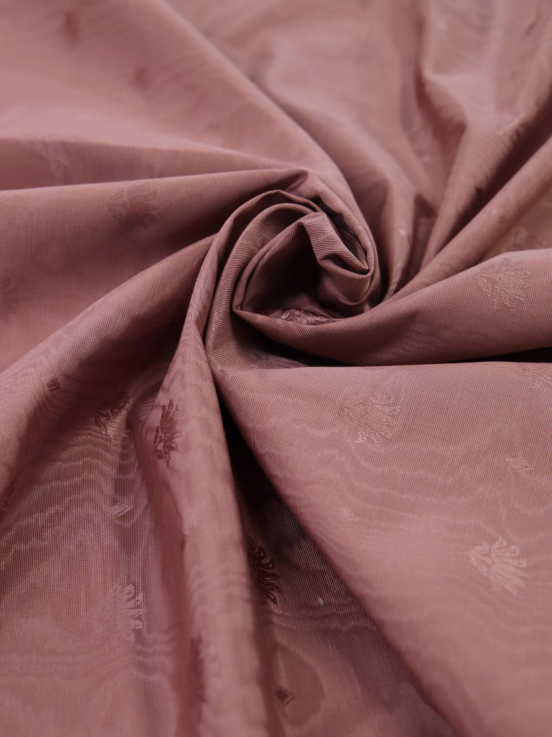May include: A dusty rose fabric featuring a subtle, repeating tonal floral pattern. The material is gathered in the center, creating soft folds and shadows that highlight the smooth texture and the delicate, woven botanical motifs throughout.
