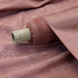May include: A bolt of dusty rose fabric featuring a subtle, tonal jacquard pattern of small floral motifs. The fabric is wrapped around a cylindrical cardboard core, showing a smooth, slightly lustrous texture with soft folds in the draped material.