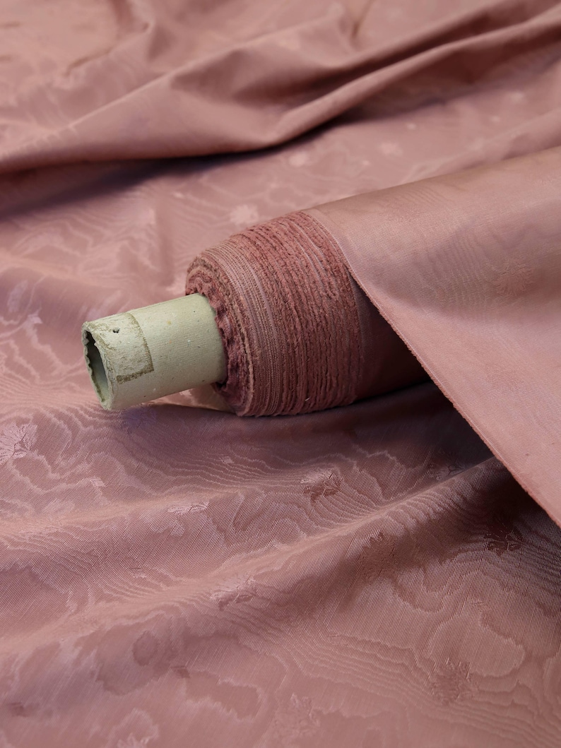 May include: A bolt of dusty rose fabric featuring a subtle, tonal jacquard pattern of small floral motifs. The fabric is wrapped around a cylindrical cardboard core, showing a smooth, slightly lustrous texture with soft folds in the draped material.