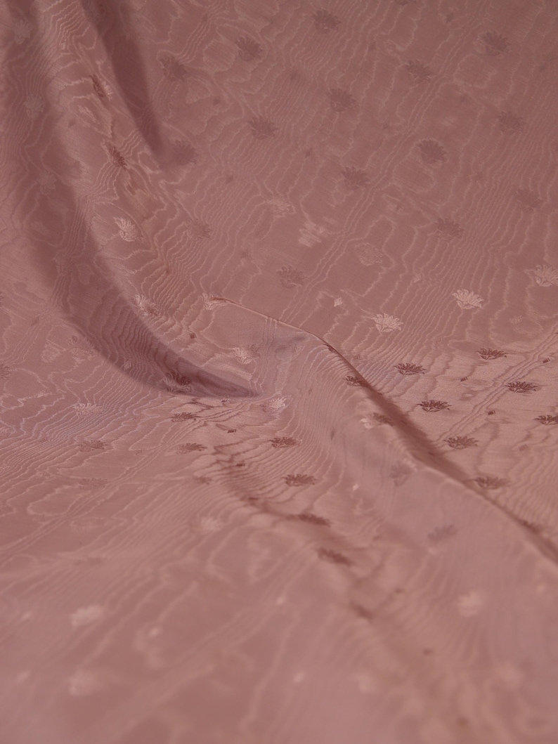 May include: A dusty rose fabric featuring a subtle, repeating floral pattern and a wavy, rippled texture. The material is draped to show soft folds, highlighting the tonal design and the consistent, muted pink color throughout the textile surface.