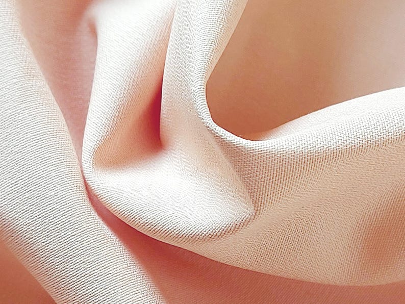 May include: Close-up of a soft, peach-coloured fabric with a smooth, silky texture. The fabric is draped and folded, creating a gentle, flowing shape.