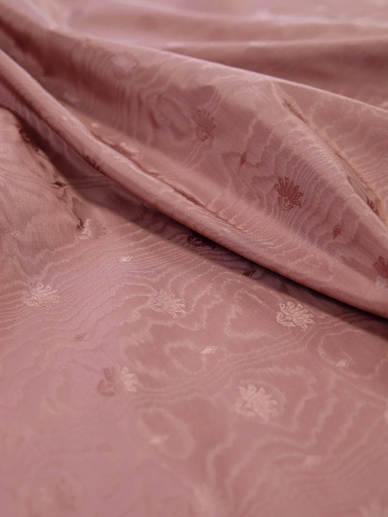 May include: A dusty rose fabric featuring a subtle, repeating floral motif and a wavy, water-like texture woven into the material. The textile is draped in soft folds, highlighting the sheen and the intricate, small-scale pattern across the surface.