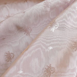 May include: A light pink fabric featuring a subtle, repeating woven pattern of small lotus-like floral motifs and tiny diamond shapes. The material has a visible, fine-ribbed texture with soft folds that create gentle highlights and shadows.