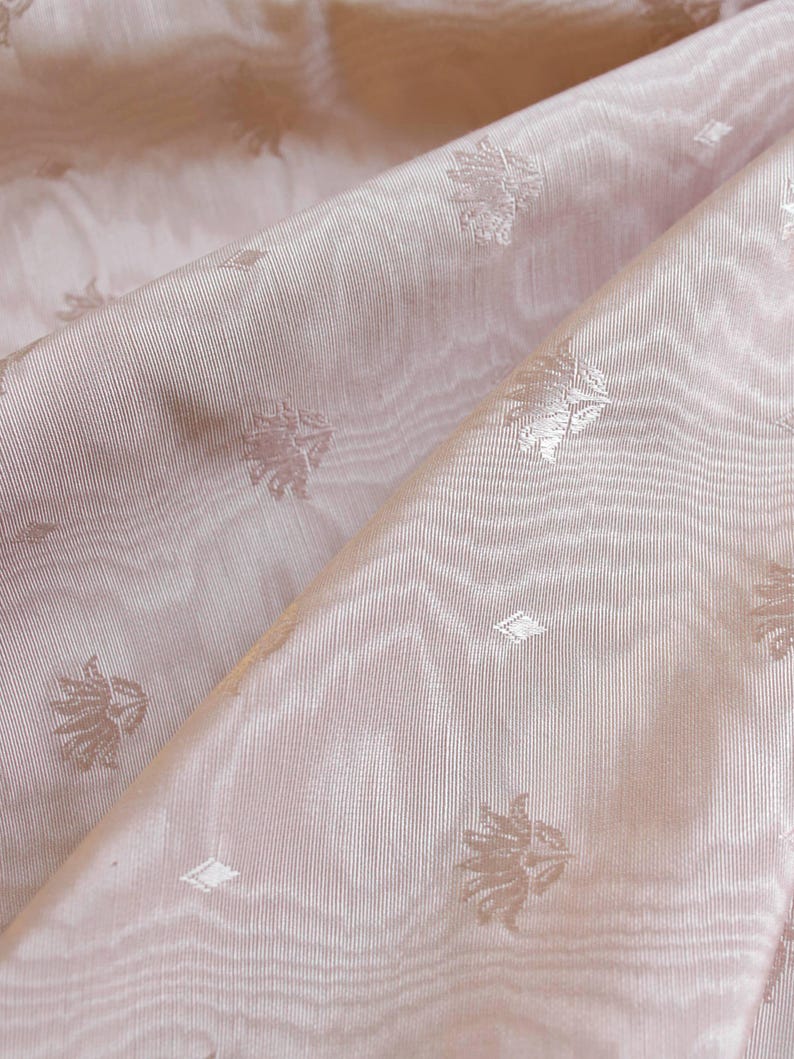 May include: A light pink fabric featuring a subtle, repeating woven pattern of small lotus-like floral motifs and tiny diamond shapes. The material has a visible, fine-ribbed texture with soft folds that create gentle highlights and shadows.