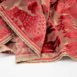 Pode incluir: Tecido de veludo vermelho com um padr&atilde;o floral dourado e vermelho. O tecido tem uma superf&iacute;cie texturizada e uma borda franjada.
