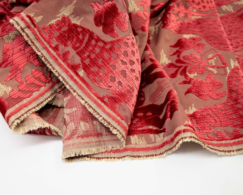 Pode incluir: Tecido de veludo vermelho com um padr&atilde;o floral dourado e vermelho. O tecido tem uma superf&iacute;cie texturizada e uma borda franjada.