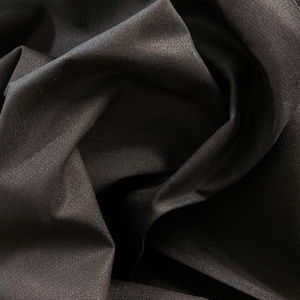 May include: Close-up of a black fabric with a textured surface. The fabric is folded and draped, showing its soft and smooth texture.
