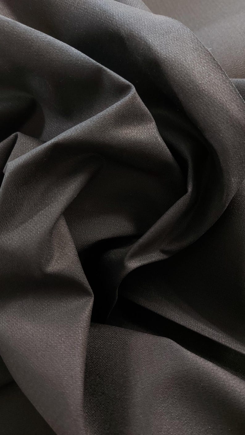 May include: Close-up of a black fabric with a textured surface. The fabric is folded and draped, showing its soft and smooth texture.