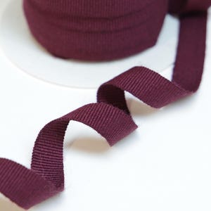 May include: A spool of burgundy-colored ribbon sits on a white surface. The ribbon is made of a woven material and is partially unraveled, with a section of the ribbon curled and lying flat. The ribbon is a solid color and has a textured appearance.