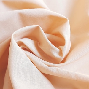 Może przedstawiać: Zbliżenie na miękką, brzoskwiniową tkaninę o gładkiej fakturze. Tkanina jest złożona i udrapowana, tworząc delikatny, płynny efekt.