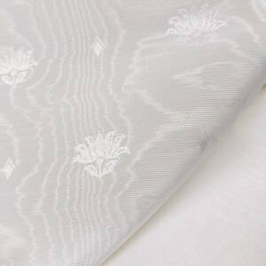 May include: A white fabric featuring a subtle, woven tonal pattern of stylized lotus flowers and small diamond shapes. The material displays a fine, diagonal ribbed texture throughout, creating a soft sheen and a layered, monochromatic visual effect.