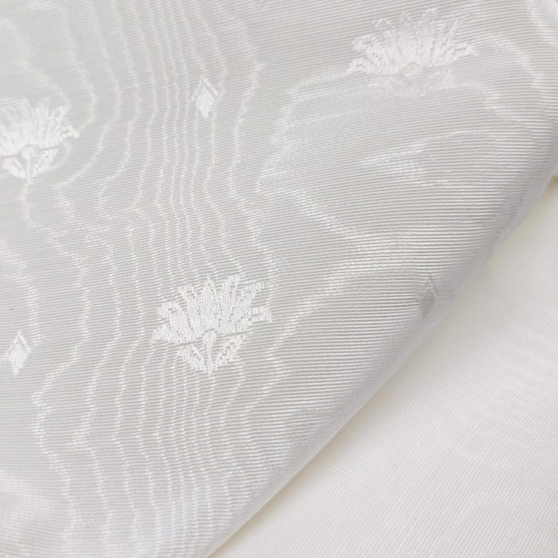 May include: A white fabric featuring a subtle, woven tonal pattern of stylized lotus flowers and small diamond shapes. The material displays a fine, diagonal ribbed texture throughout, creating a soft sheen and a layered, monochromatic visual effect.