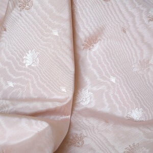 May include: A pale pink fabric featuring a subtle, repeating woven pattern of small floral motifs and tiny diamond shapes. The material has a soft, lustrous sheen with visible diagonal ridges and folds that create depth across the surface.