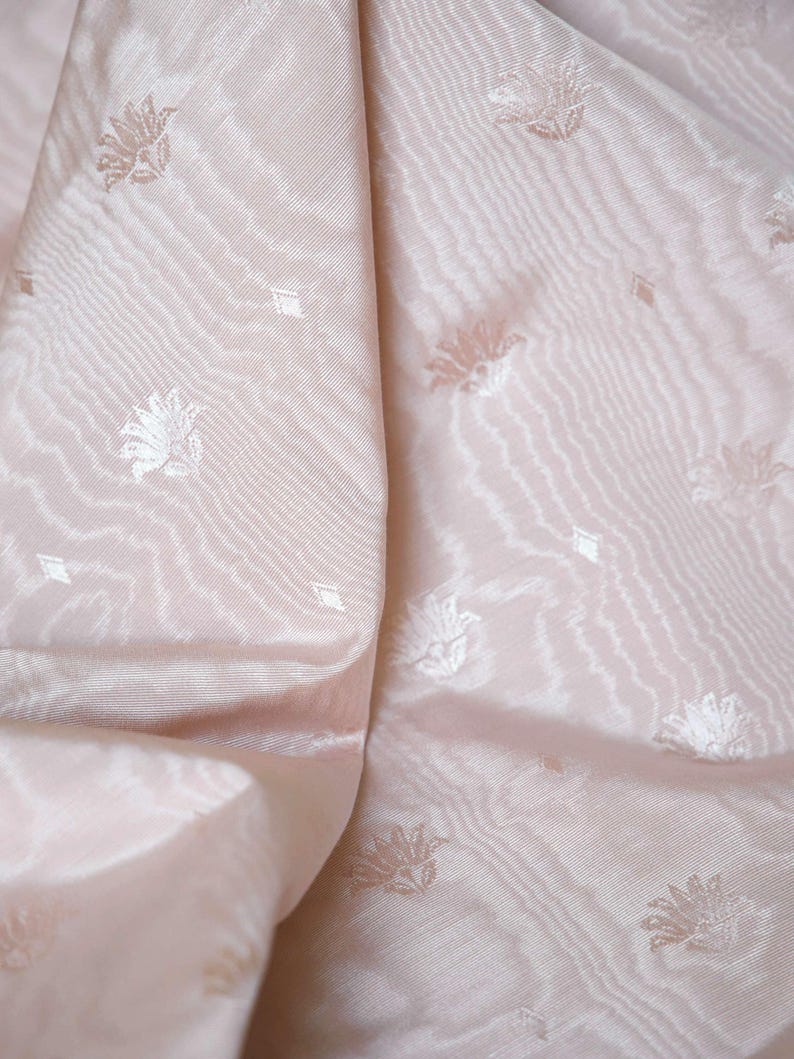 May include: A pale pink fabric featuring a subtle, repeating woven pattern of small floral motifs and tiny diamond shapes. The material has a soft, lustrous sheen with visible diagonal ridges and folds that create depth across the surface.