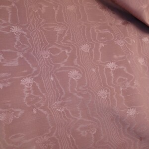May include: A dusty rose fabric featuring a subtle, tonal jacquard pattern of small, stylized lotus-like flowers arranged in vertical columns. The textile displays a delicate, wavy, wood-grain-like texture that runs parallel to the floral motifs.