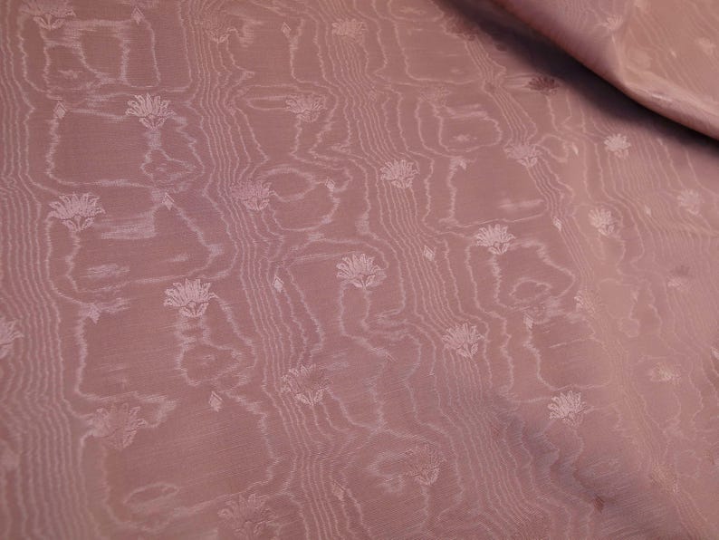 May include: A dusty rose fabric featuring a subtle, tonal jacquard pattern of small, stylized lotus-like flowers arranged in vertical columns. The textile displays a delicate, wavy, wood-grain-like texture that runs parallel to the floral motifs.