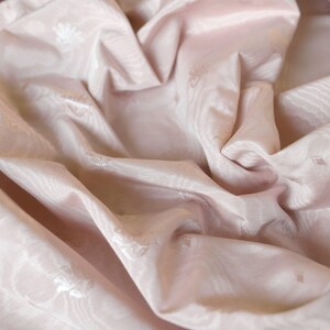 May include: A pale pink fabric featuring a subtle, tonal jacquard pattern of small floral motifs and geometric shapes. The material has a smooth, slightly lustrous finish with deep, soft folds that create a play of light and shadow across the surface.
