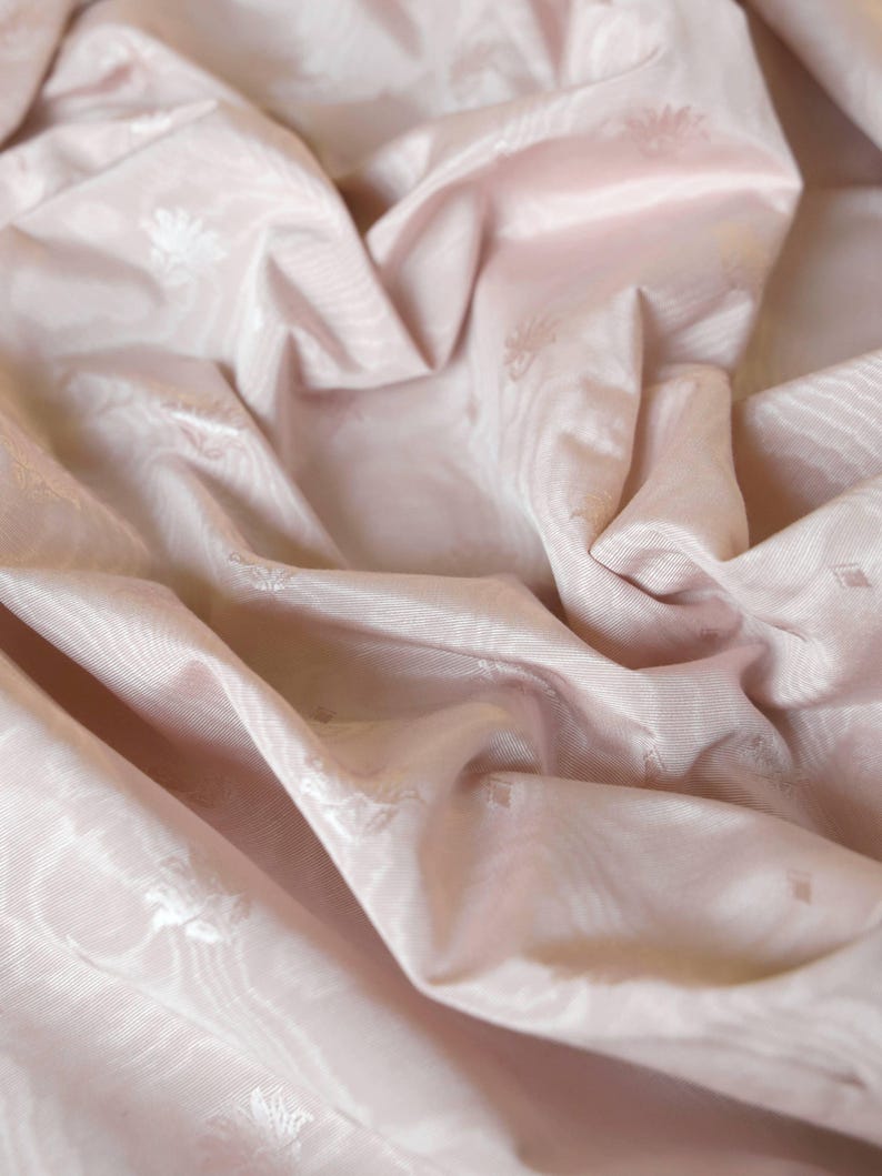 May include: A pale pink fabric featuring a subtle, tonal jacquard pattern of small floral motifs and geometric shapes. The material has a smooth, slightly lustrous finish with deep, soft folds that create a play of light and shadow across the surface.