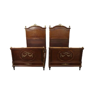 May include: Two antique wooden beds with ornate gold-colored accents. Each bed features a headboard with a woven panel and a footboard with an oval medallion. The beds have decorative trim and stand on short legs.