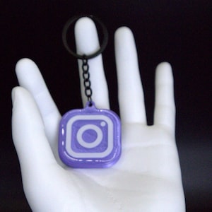 May include: A purple and white Instagram logo keychain. The keychain features a square design with rounded corners and a white Instagram logo. It hangs from a black chain and keyring. The background is black.