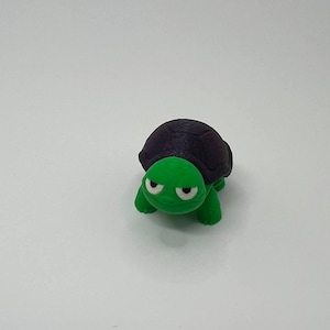 Peut inclure: Une petite figurine de tortue en plastique verte et noire avec une expression boudeuse. La tortue a une carapace noire et des pattes et une tête vertes.
