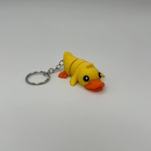 Puede incluir: Un llavero de pato de goma amarillo con un anillo de llave plateado. El pato tiene ojos negros y un pico naranja.
