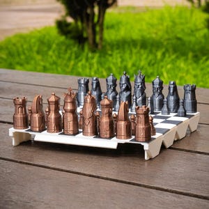 Op de afbeelding: Een compleet schaakspel met bronzen en grijze stukken op een zwart-wit geblokt bord. De schaakstukken zijn gerangschikt op een witte basis, geplaatst op een houten tafel met een groene achtergrond.