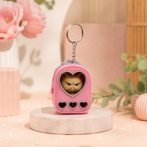 May include: A pink miniature backpack keychain featuring a heart-shaped window on the front. Inside the window, a small, grumpy-looking tan cat figurine is visible. The backpack has three small heart-shaped cutouts at the bottom and a silver ring.