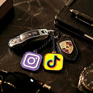May include: A black Porsche car key fob attached to a metal ring with three charms: a purple Instagram logo square, a yellow TikTok logo square, and a black leather shield-shaped tag featuring the colorful Porsche crest logo.