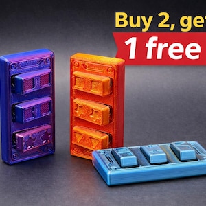 Könnte beinhalten: Drei 3D-gedruckte elektronische Komponenten in Blau, Orange und Lila. Die Komponenten haben eine rechteckige Form mit mehreren rechteckigen Tasten. Der Text "Buy 2, get 1 free" befindet sich in der oberen rechten Ecke.