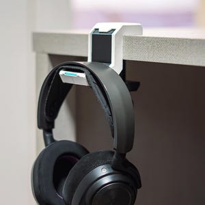 May include: A black over-ear headset with plush ear cushions and a wide headband, suspended from a white and black clamp-style headphone hanger. The hanger is secured to the edge of a flat surface, featuring a small teal accent on the inner hook.