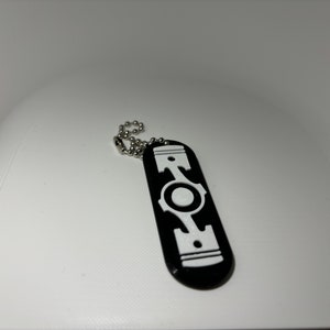 May include: Black and white keychain with a piston design. The keychain is made of metal and has a silver chain.