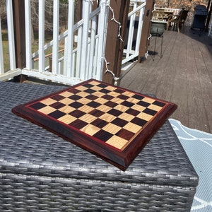 May include: A wooden chessboard with a dark brown frame and a red accent border. The squares alternate between light and dark wood tones. The board is set on a gray woven surface, with a deck and trees in the background.