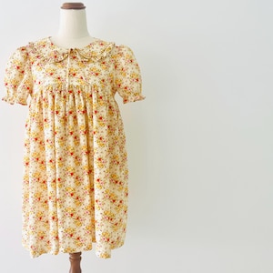 Könnte beinhalten: Ein cremefarbenes Kleid mit einem lebendigen Blumenmuster in Rot-, Gelb- und Grüntönen. Das Kleidungsstück verfügt über kurze Puffärmel mit gerüschten Kanten, einen runden Kragen mit Rüschenbesatz und eine Silhouette mit geraffter Taille.